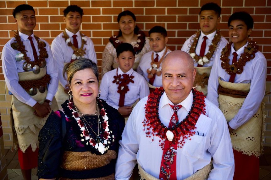 Tevita and Sharon Ta'ai and nine of their 13 kids pose in traditional dress in front of a brick wall.