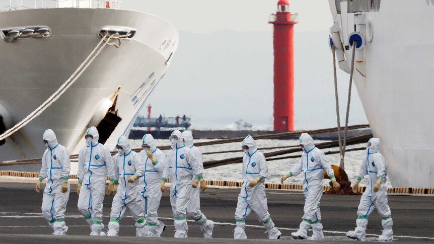 People in white hazmat suits walking across a dock with a ship beside them