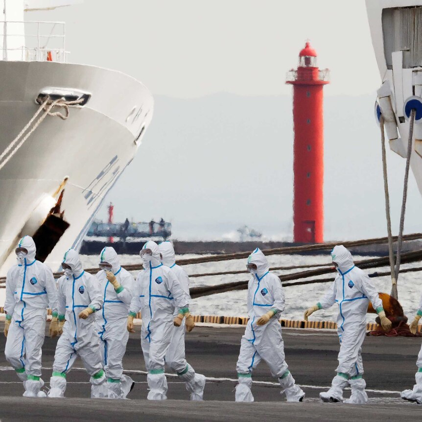 People in white hazmat suits walking across a dock with a ship beside them