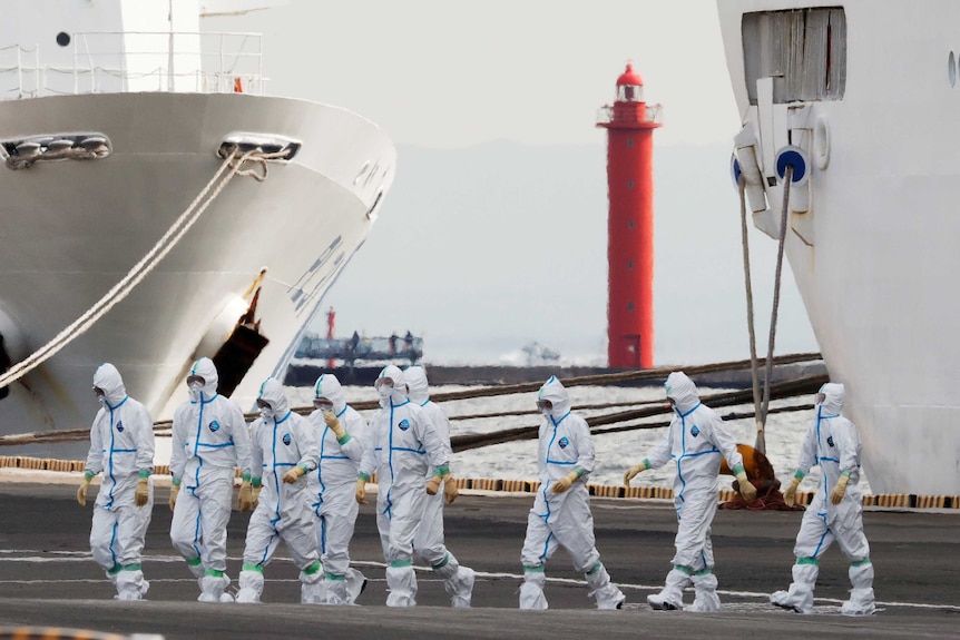 People in white hazmat suits walking across a dock with a ship beside them