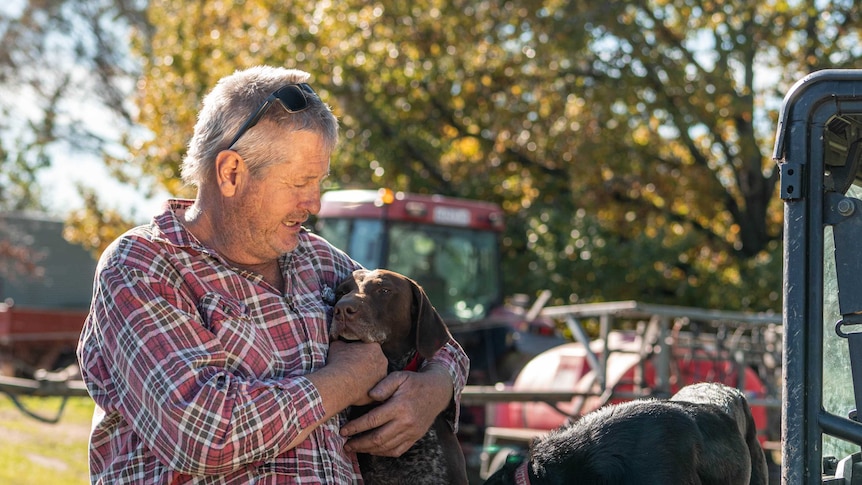 A man with grey hair, wearing a flannelette shirt, hugs his dog which is sitting on the back of a farm vehicle.