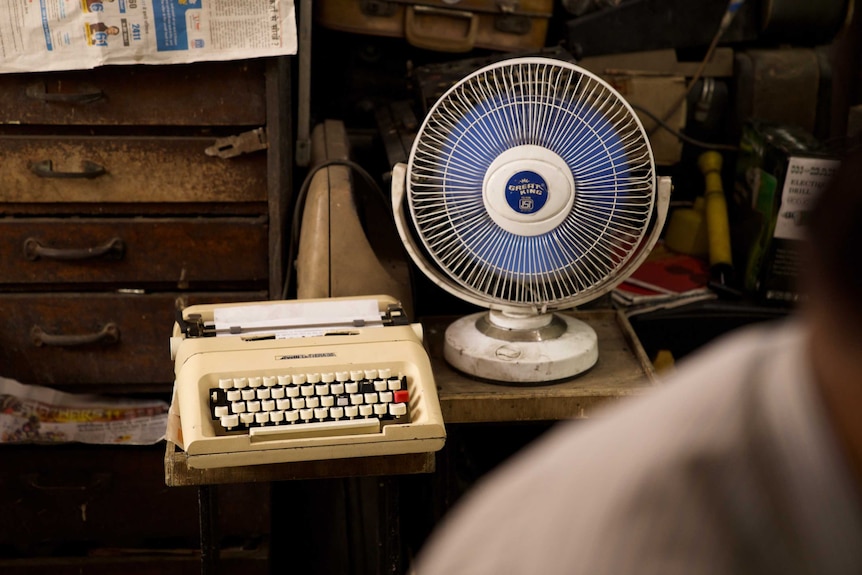 One of the more modern typewriters  on a small table next to a desktop fan