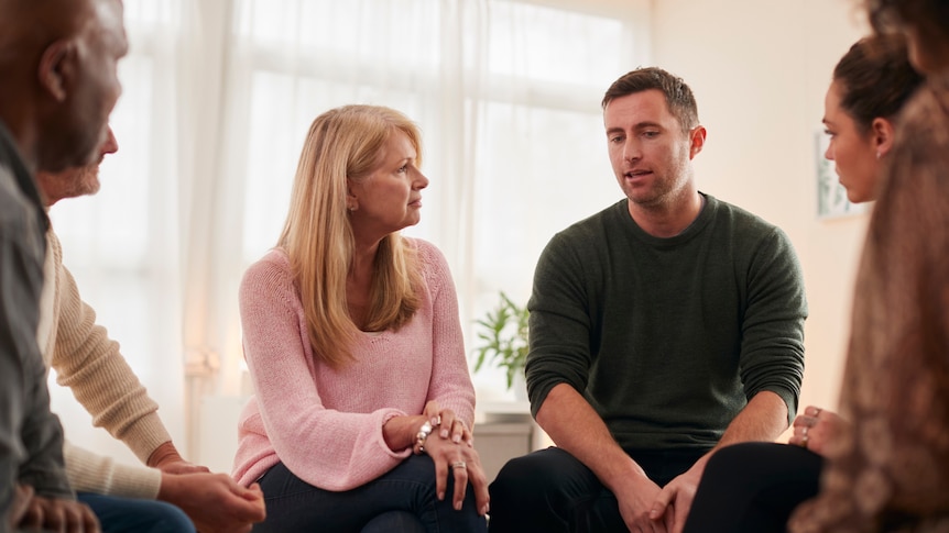 man with short hair sitting on a stool talking to other adults