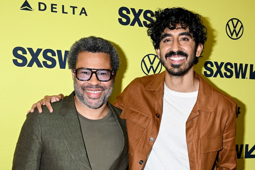 A black man wearing a suit and glasses and a man of South Asian heritage stand in front of a yellow background
