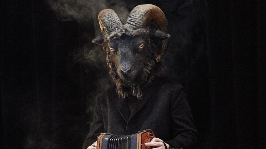 A man wearing a goat mask sits on stage playing a concertina.