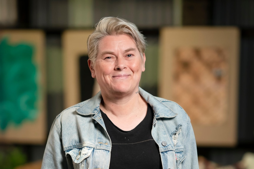 White middle-aged woman with short blonde hair wears black shirt and pale blue denim jacket on a TV set.