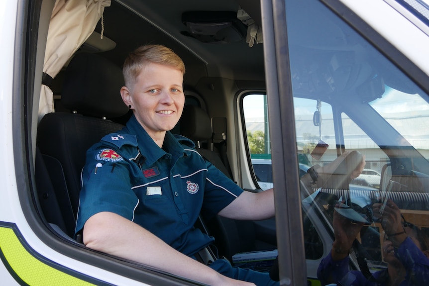 A woman with blonde short hair sits in the driver's seat of an ambulance and looks out the window smiling.
