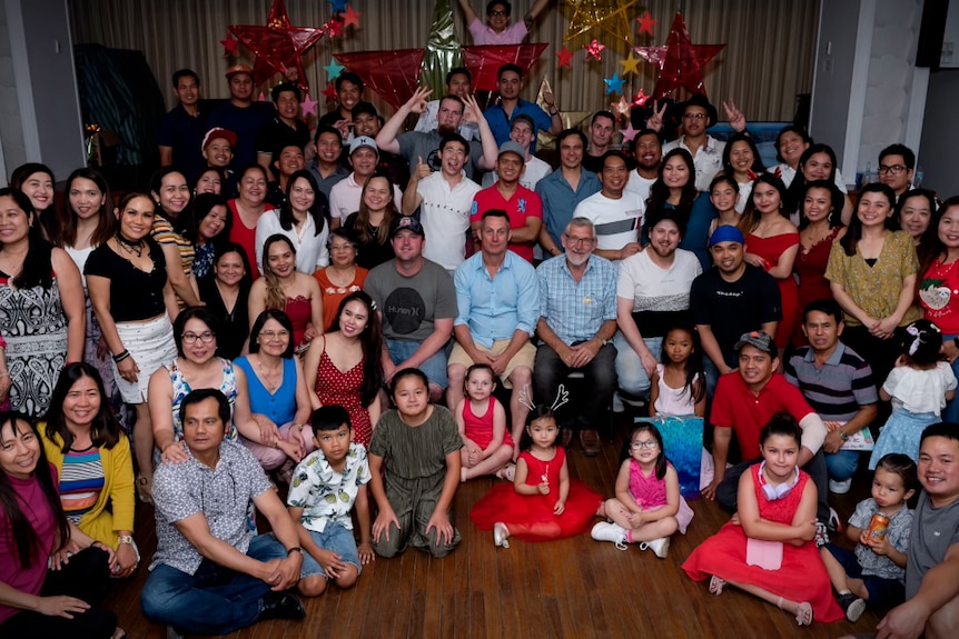 Large group of people of multiple nationalities grouped together in a community hall. Christmas decorations in the background.