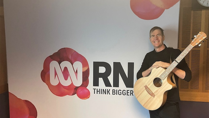 Man stands smiling in front of RN Breakfast photo wall with a guitar over his chest 