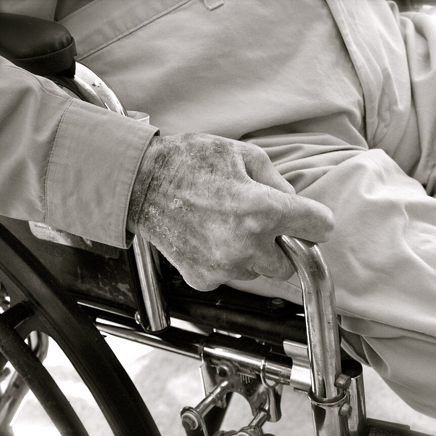 Older man in sitting in a wheel chair. You can see his legs and one hand which is holding the arm rest.