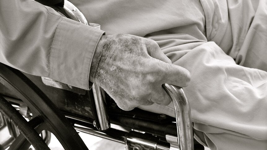 Older man in sitting in a wheel chair. You can see his legs and one hand which is holding the arm rest.