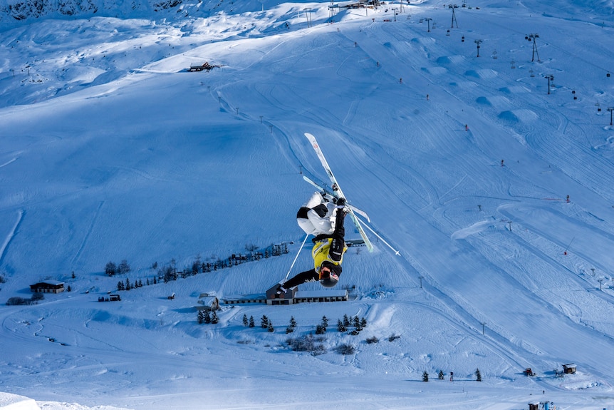 Jakara Anthony jumps wearing skis upside down in front of a white snowy backdrop