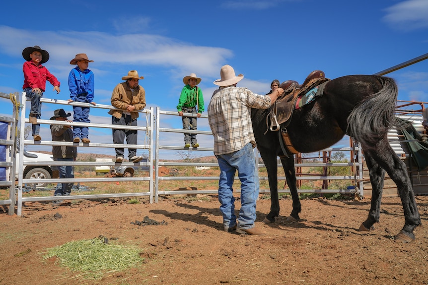 A group of young boys and men dressed in stockmen gear stand around a man with a horse.