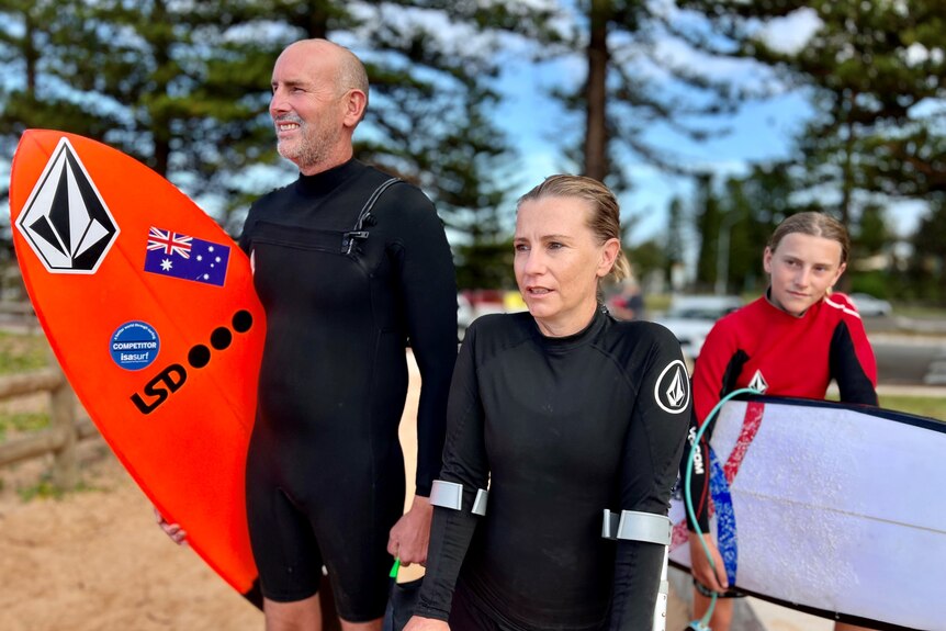 A man, woman and teenage girl look at the ocean while wearing wetsuits.