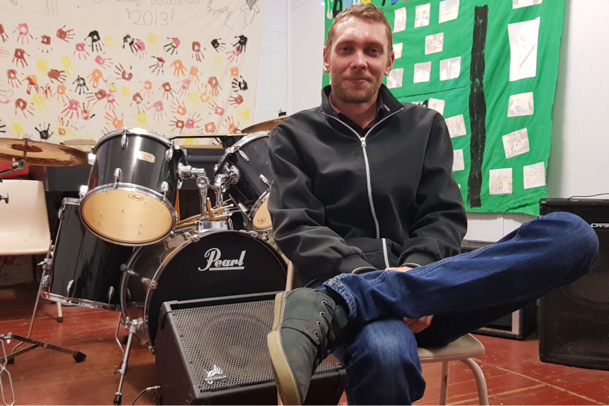 A man wearing a black jacket and blue jeans sits on a chair in front of a drum kit.