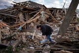 A woman bending to water a few flowers amidst the rubble of her home.