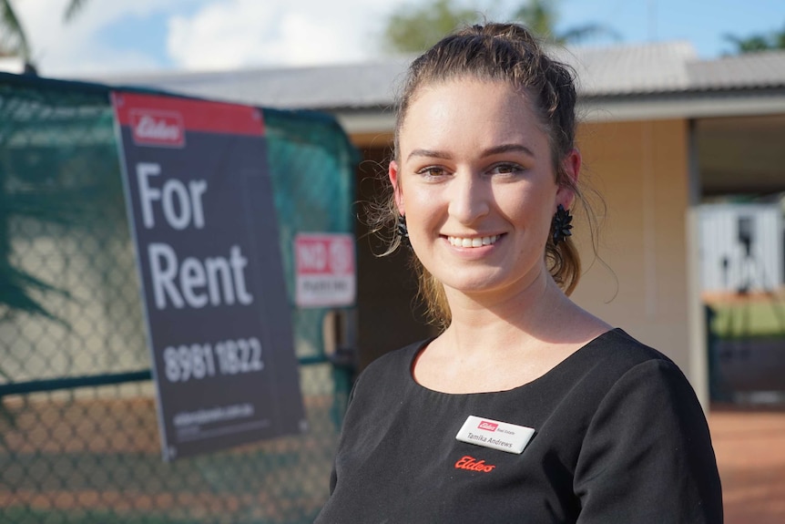 A young woman smiles at the camera with a for rent sign in the background.