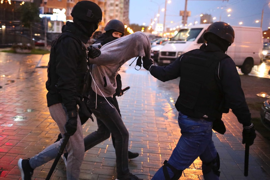 Three police in black and helmets drag a person by their grey hoodie.