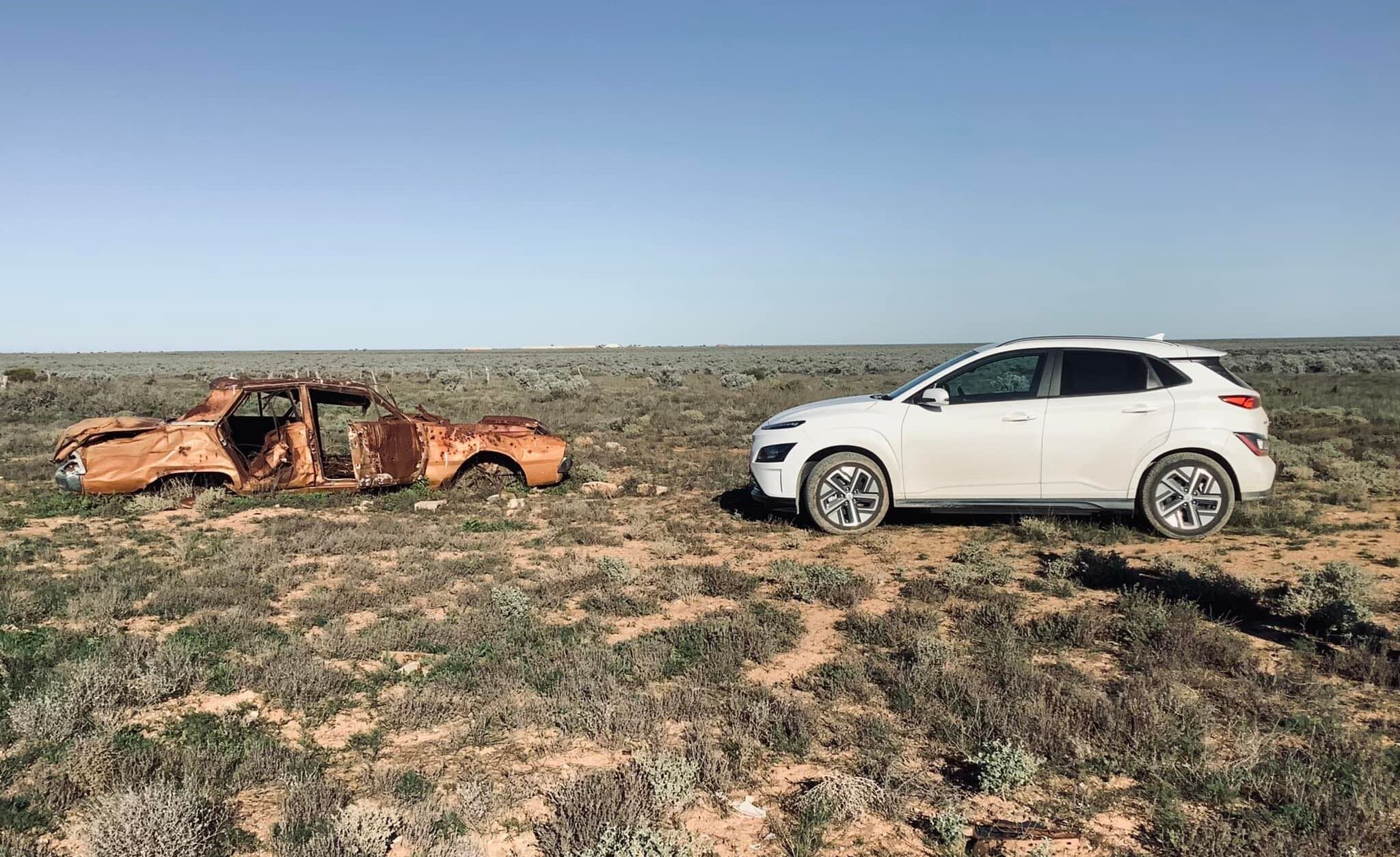 A white electric vehicle facing an old, abandoned car on the Nullarbor Plains