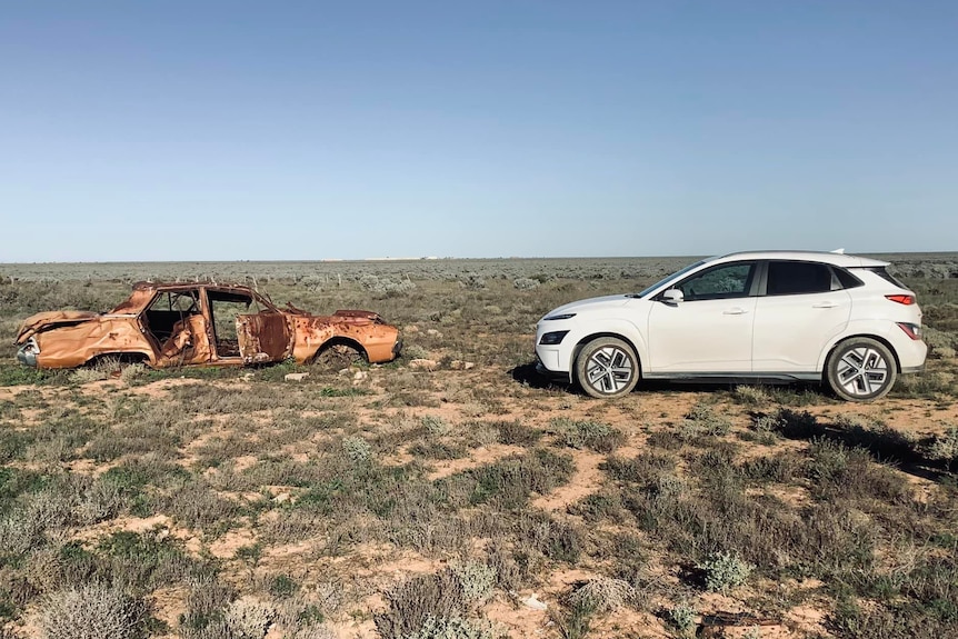 A white electric vehicle facing an old, abandoned car on the Nullarbor Plains