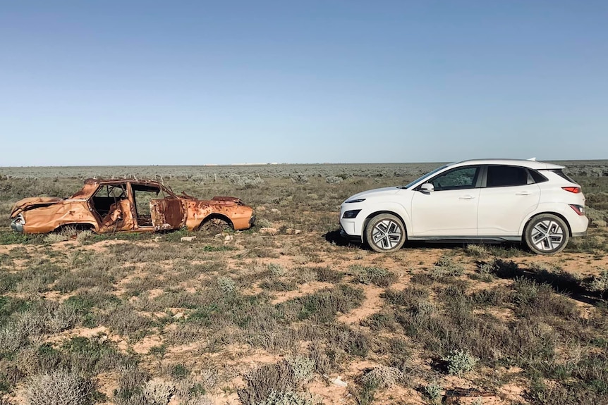 Un véhicule électrique blanc face à une vieille voiture abandonnée sur les plaines de Nullarbor