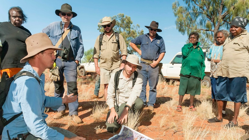 Group of people standing and crouching around a map on red dirt