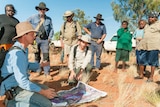 Group of people standing and crouching around a map on red dirt