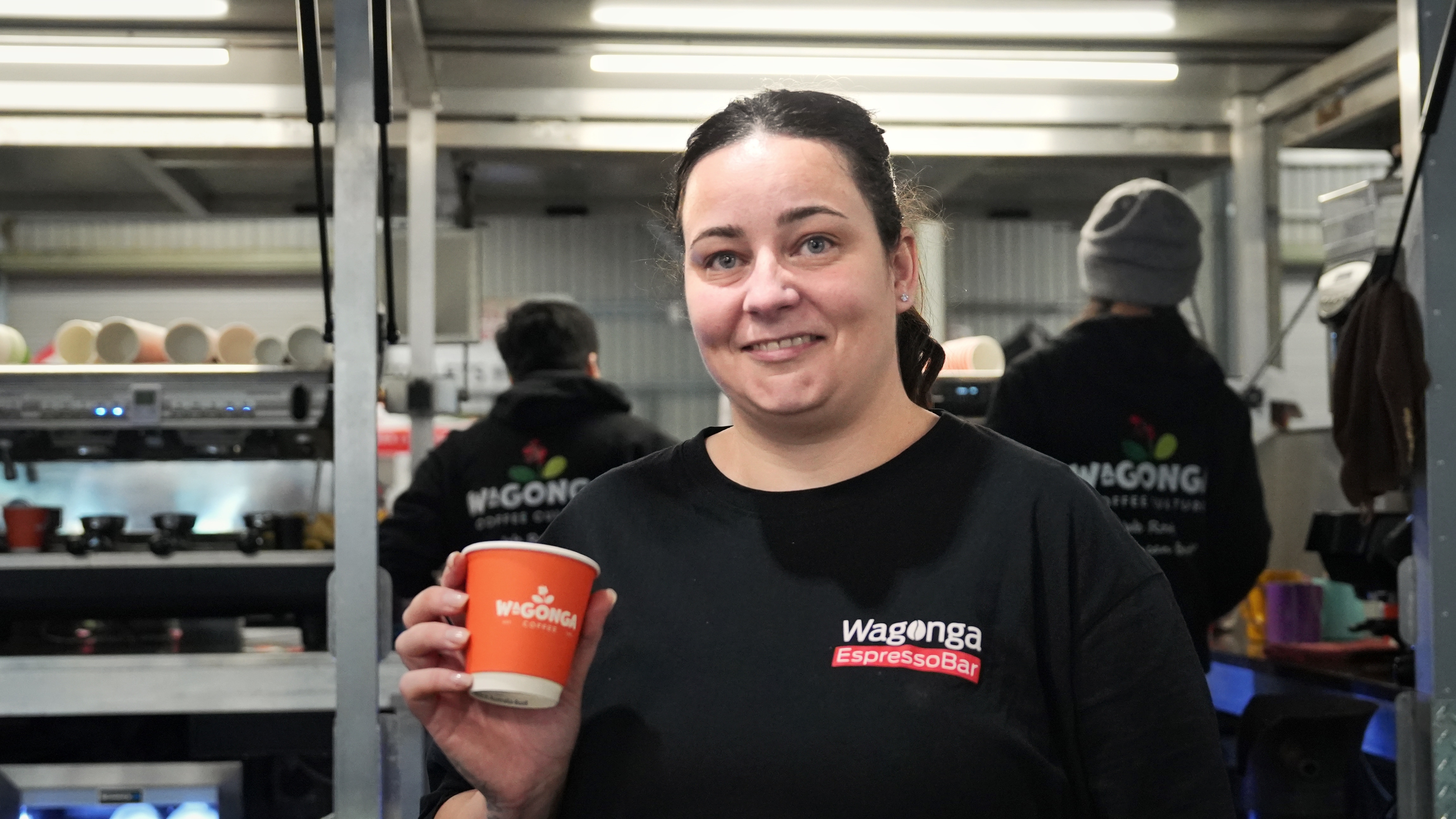 A woman with dark hair pulled into a ponytail holds up an orange paper coffee cup.