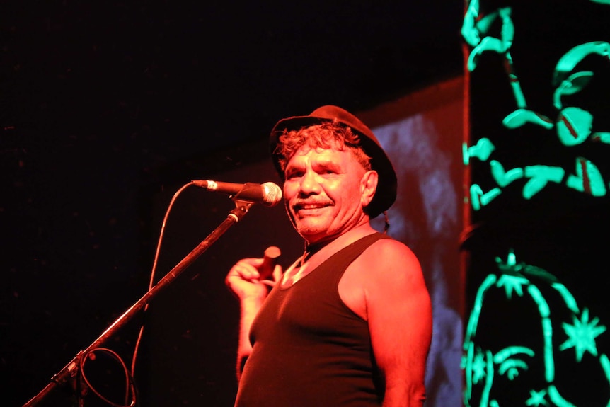 Aboriginal man on stage under a red light smiling with a black hat on.