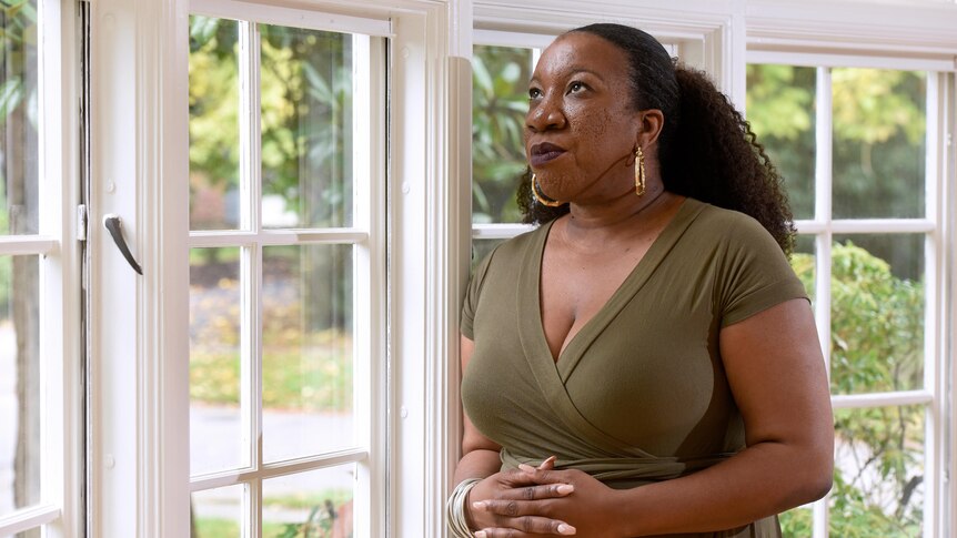 A portrait of Tarana Burke standing by a window in her home. 