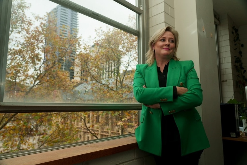 A blonde woman in a green blazer smiling with her arms folded, with city buildings in the window behind her.