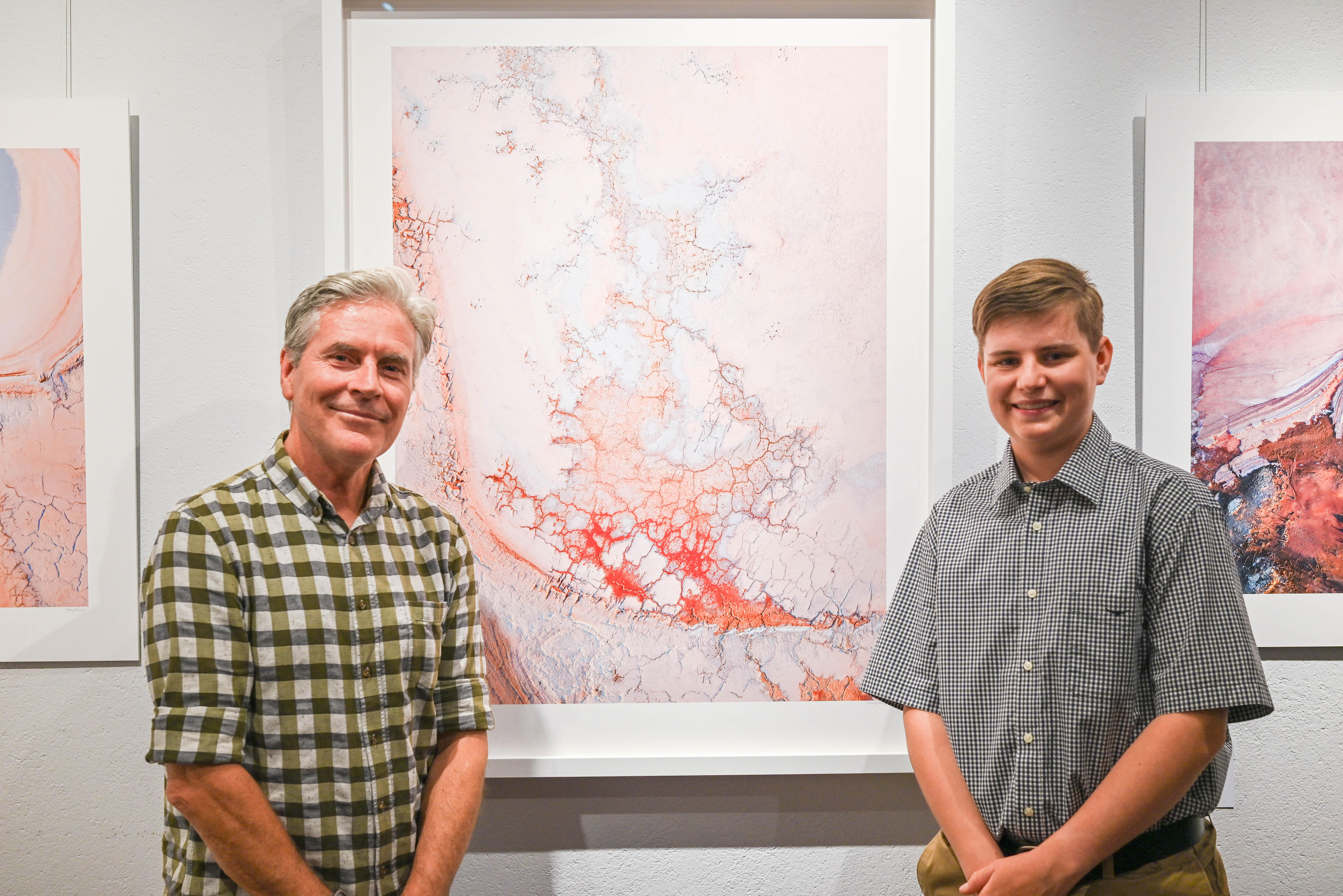 A middle-aged, silver-haired man and a young man with golden-brown hair standing near photographs hanging in a gallery.