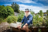 A man with a white beard sits next to a vegetable patch