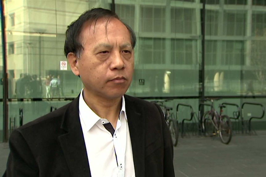 Qin Jin wears a white shirt with no tie and black blazer as he speaks out the front of a large glass building.