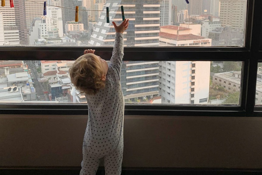 A child in a onesie grabs at a peg on a clothes line as he leans on a closed window overlooking the city.