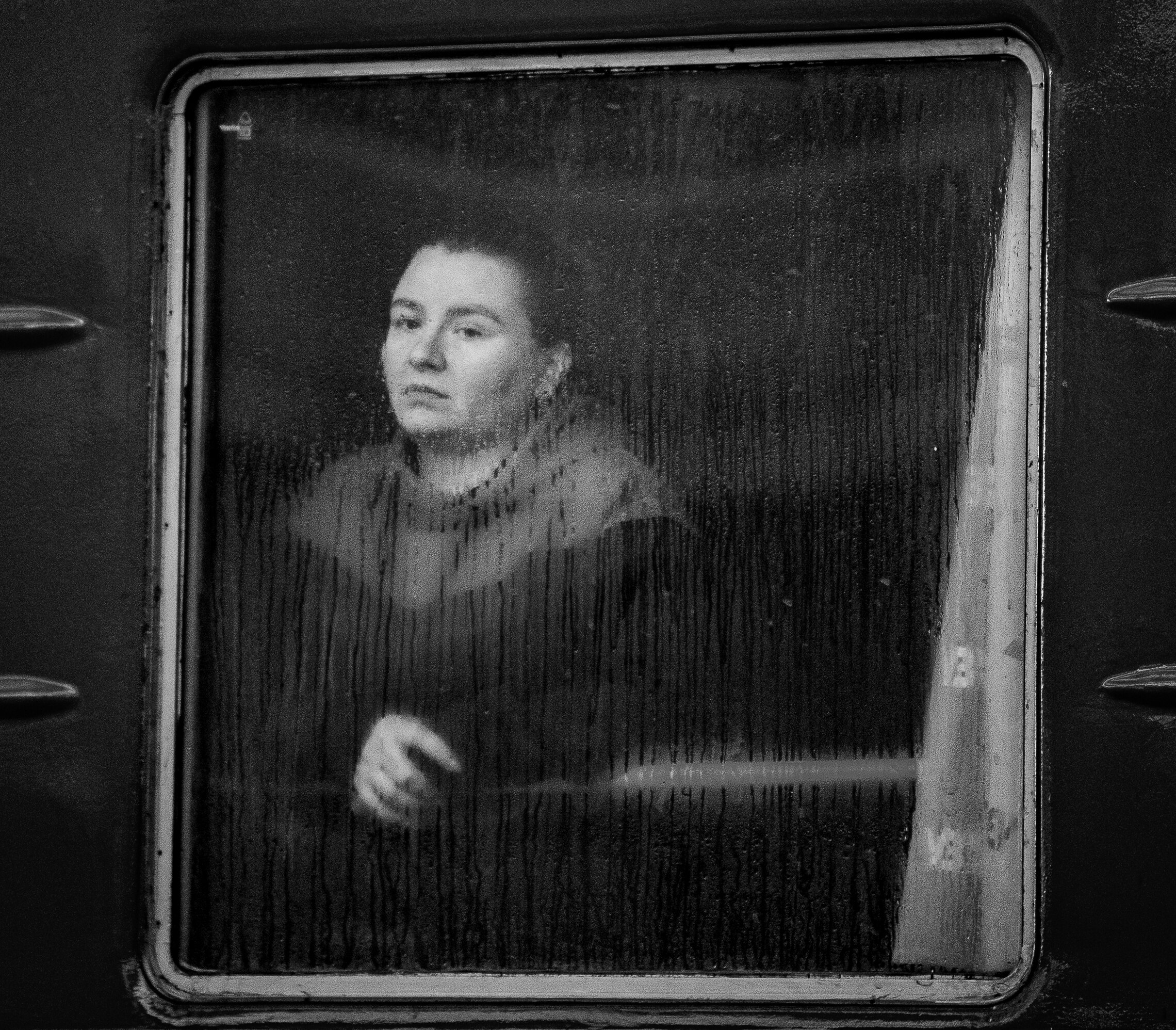 Black and white photo of woman looking sadly out of a window with rain drops falling on glass.