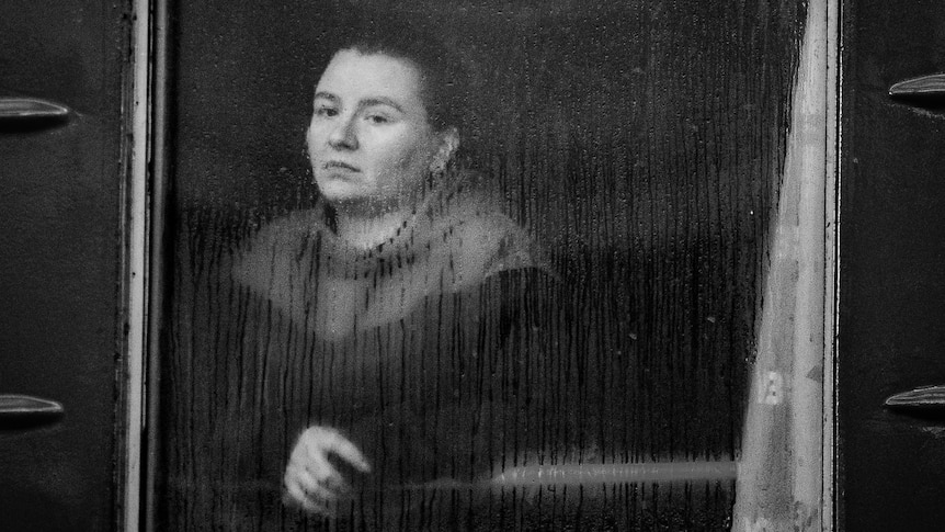 Black and white photo of woman looking sadly out of a window with rain drops falling on glass.
