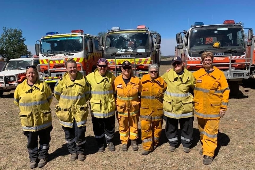 Seven women in various shades of yellow bushfire protective gear stand arm-in-arm in front of three large fire fighting tankers