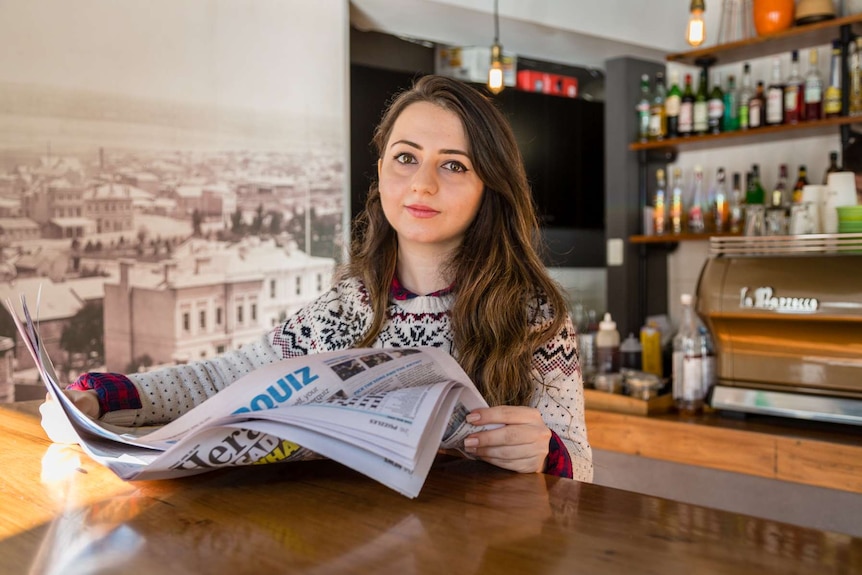Iraqi refugee Marwa Albana sits in a coffee shop, she is holding a newspaper and looking at the camera.