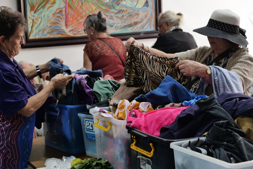Women stand around large tubs filled with clothes on a table, picking up items to examine them.
