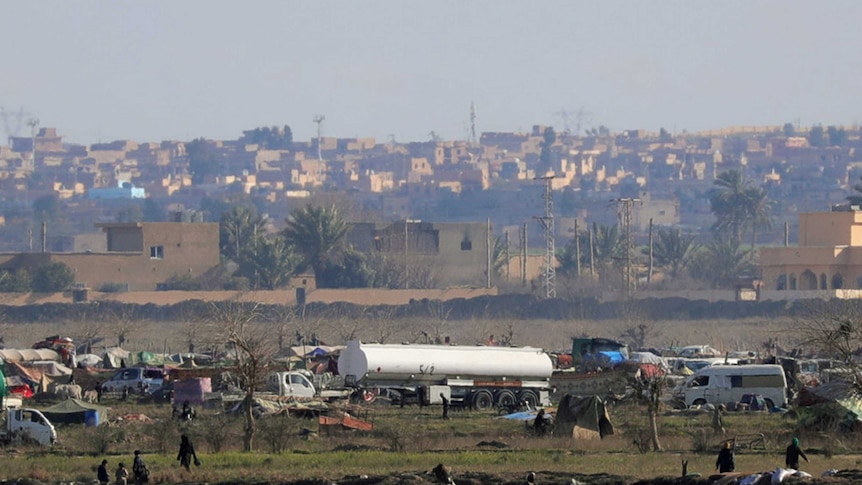 A distant photo of a series of tents and vehicles with a dusty village visible in the background.