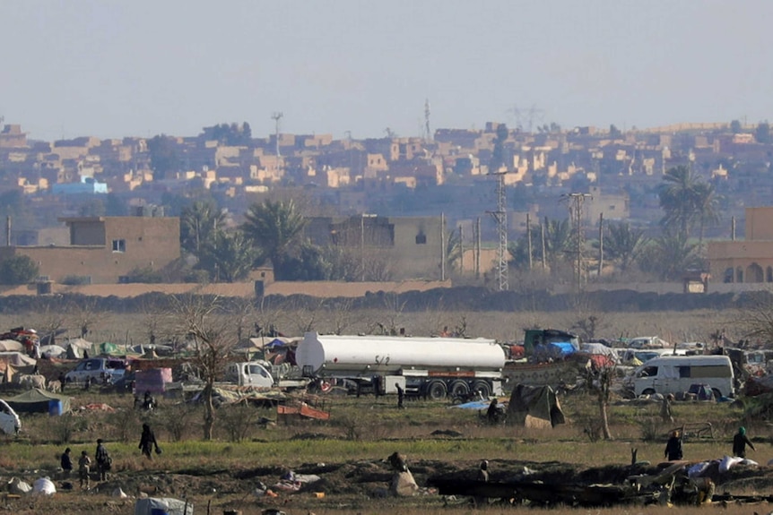 A distant photo of a series of tents and vehicles with a dusty village visible in the background.