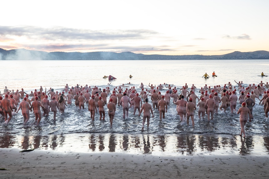 A group pf naked people wearing swimming caps standing in the water at a beach.