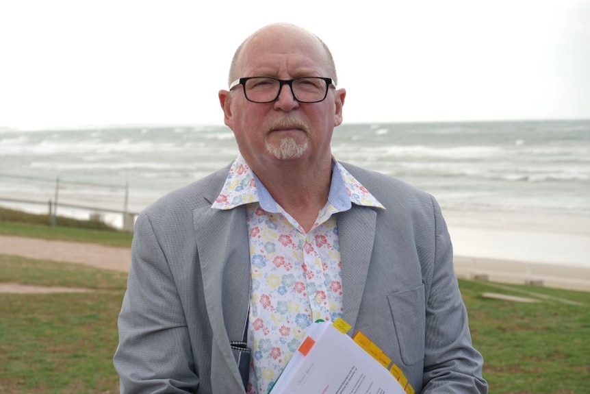 A man wearing black glasses, grey blazer and floral shirt stands on a lawn in front of the ocean.