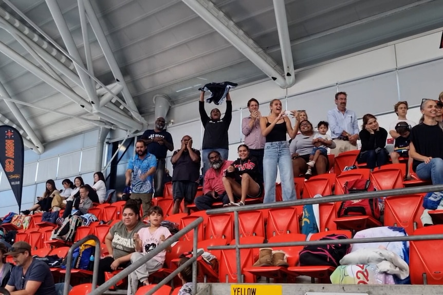 A group of adults cheers from seating at the top of a swimming centre stadium.