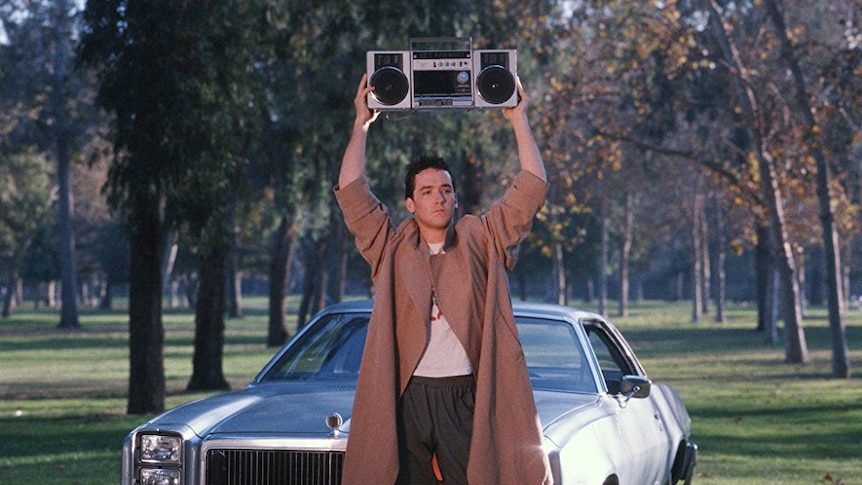 John Cusack holds a boombox above his head in front of his car.