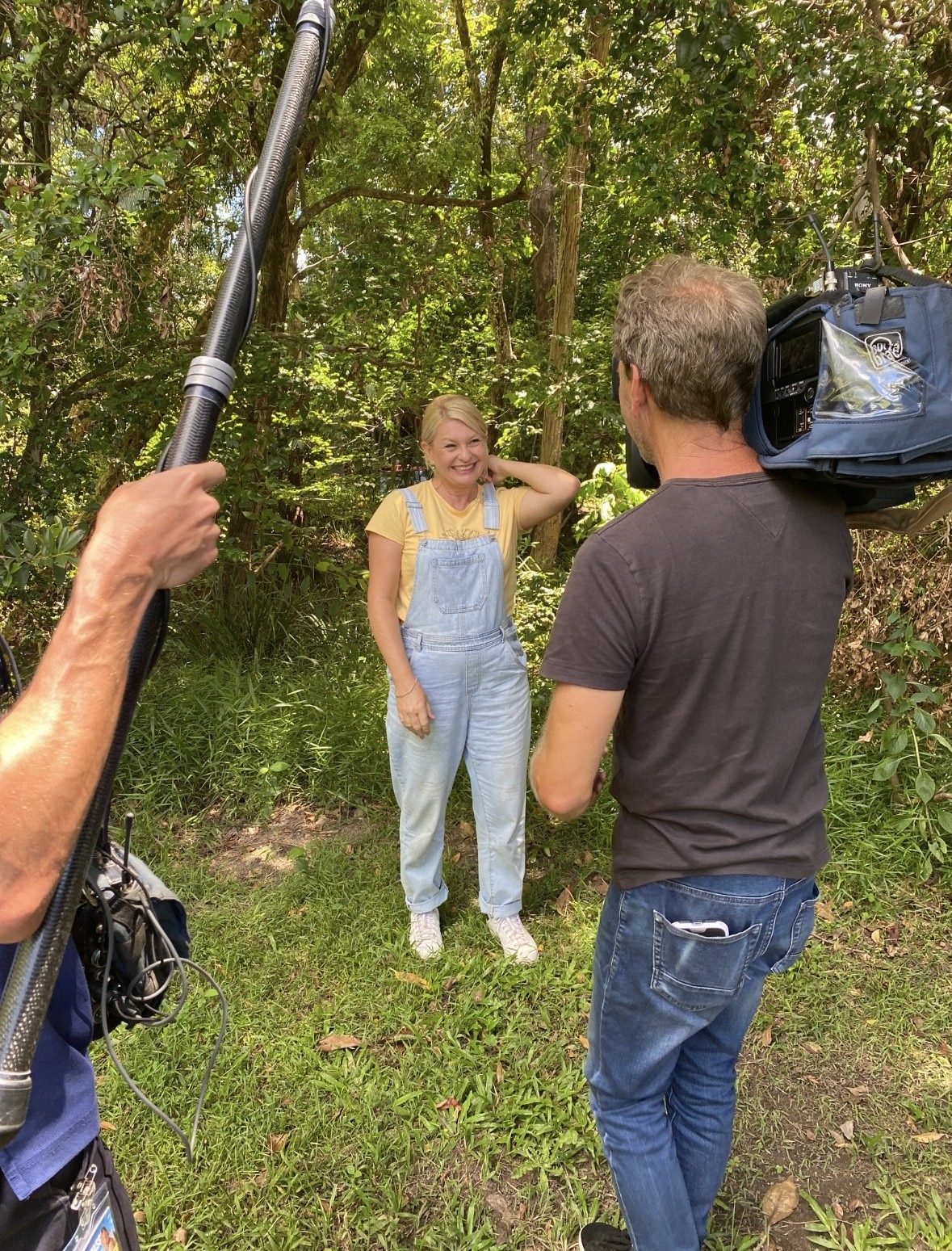 A smiling blonde woman in denim overalls is being filmed by a TV cameraman with a boom microphone held above.