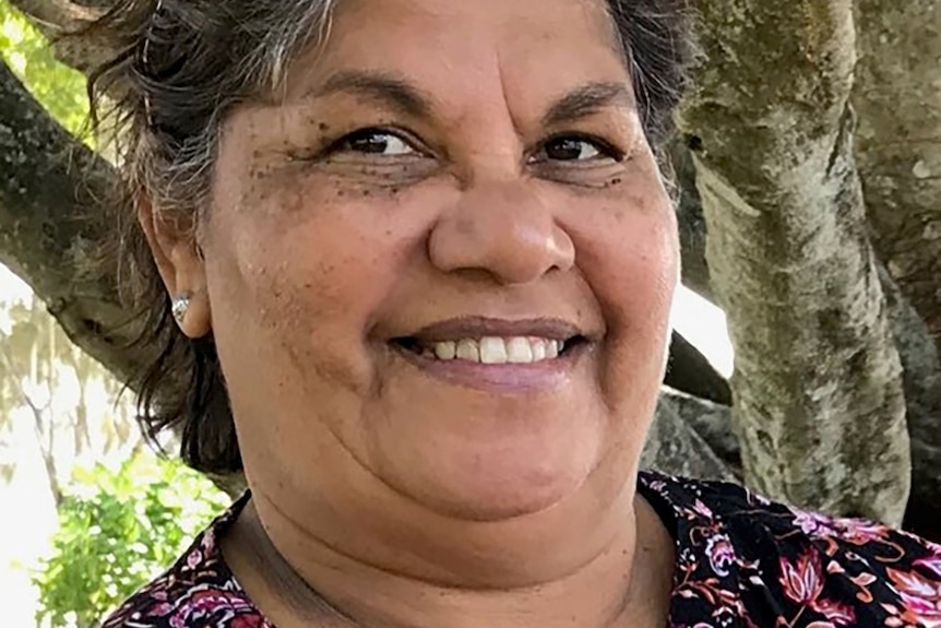 A close up of a smiling older woman, salt and pepper hair, black and pink printed top in front of a tree.