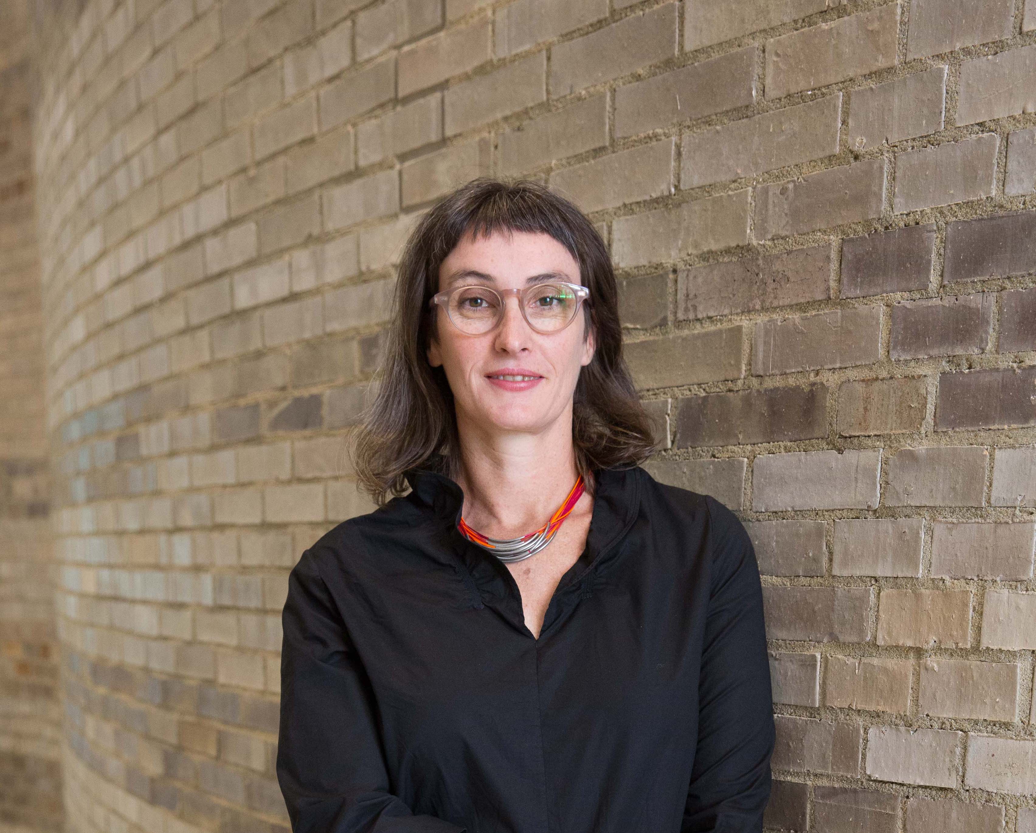 A female professor with shoulder-length brown hair and glasses is standing against a brick wall.