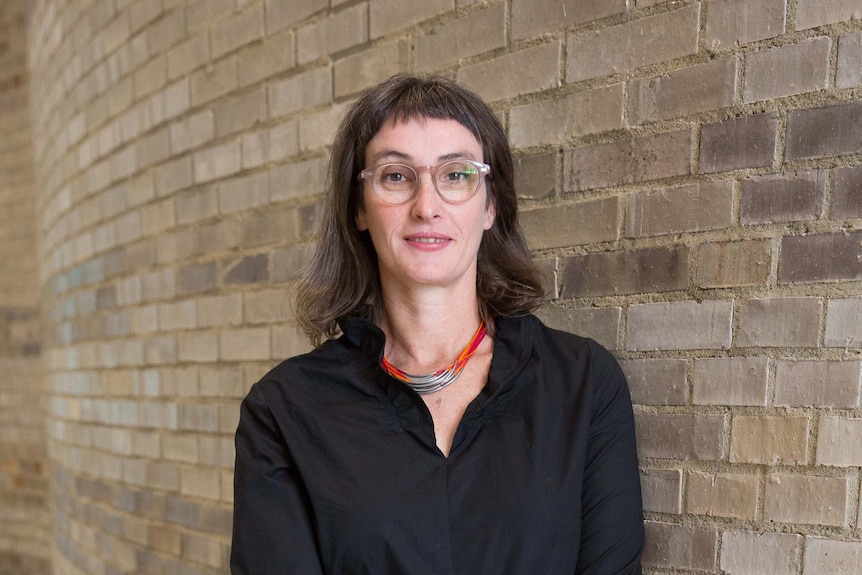 A female professor with shoulder-length brown hair and glasses is standing against a brick wall.